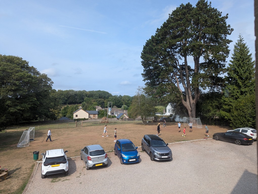Wide photo, view from the upper large hall, of several campers playing football beyond parked cars. The scene includes some large trees and a tree-lined horizon.