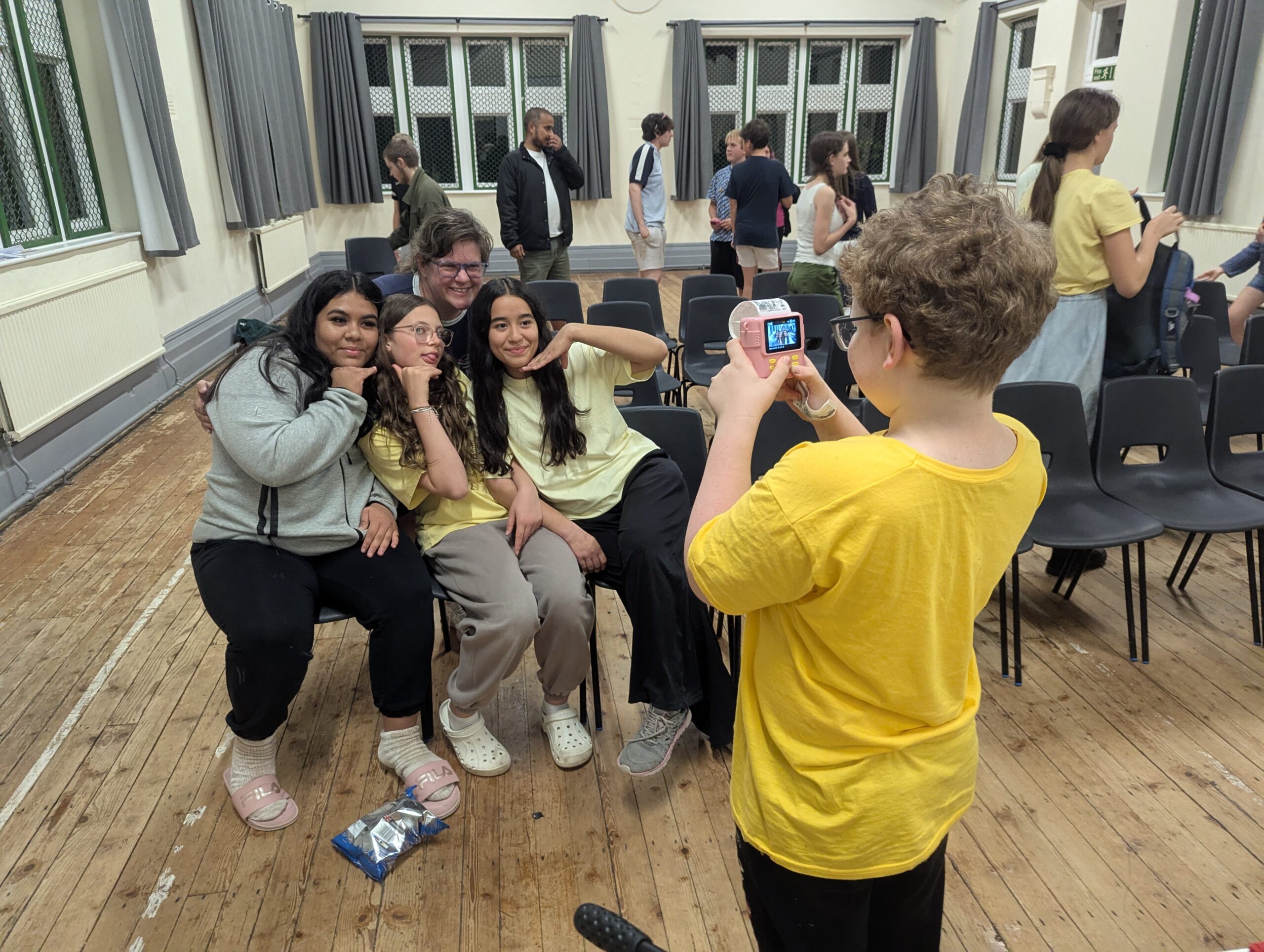 3 Christian Worship Campers in the upper hall of Woodcroft posing for a photo being taken by a young camper with a pink thermal paper printing camera. A female leader poses behind. Other campers are milling about in the background, some moving chairs.