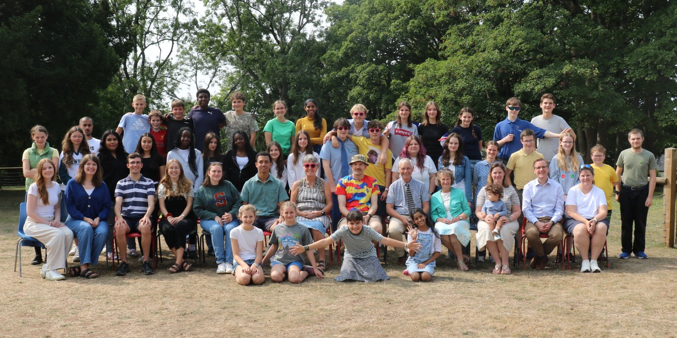 With brown (dry) grass and trees behind, four rows of people of all ages in a portrait - Chepstow Woodcroft in August 2025