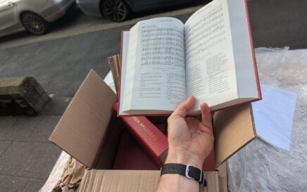 Background is a street pavement with parked cars and in the foreground an opened box of new red hymn books, with a hand and forearm holding one of the books open on number 289, Great is the Gospel of our Glorious God.