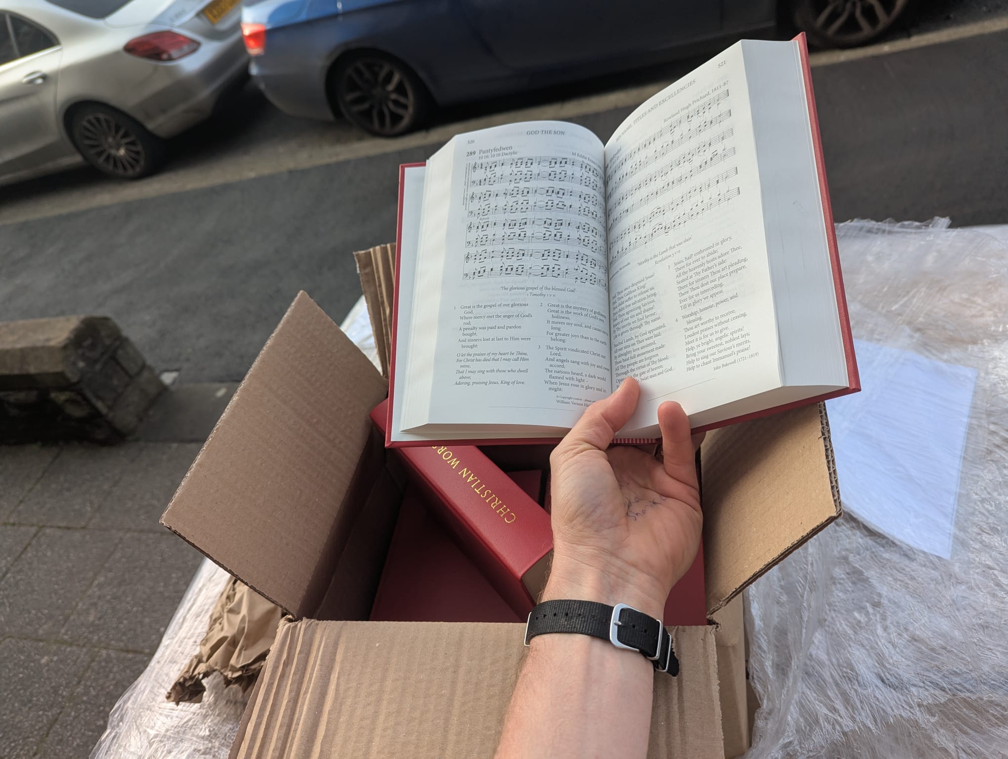 Background is a street pavement with parked cars and in the foreground an opened box of new red hymn books, with a hand and forearm holding one of the books open on number 289, Great is the Gospel of our Glorious God.