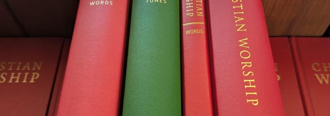 Four hymnbooks sit on their edges atop of a pile of red hymnbooks. The newest 'Combined Words and Music' edition is the widest and furthest to the right