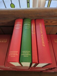 Four hymnbooks sit on their edges atop of a pile of red hymnbooks. The newest 'Combined Words and Music' edition is the widest and furthest to the right