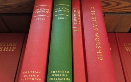 Four hymnbooks sit on their edges atop of a pile of red hymnbooks. The newest 'Combined Words and Music' edition is the widest and furthest to the right