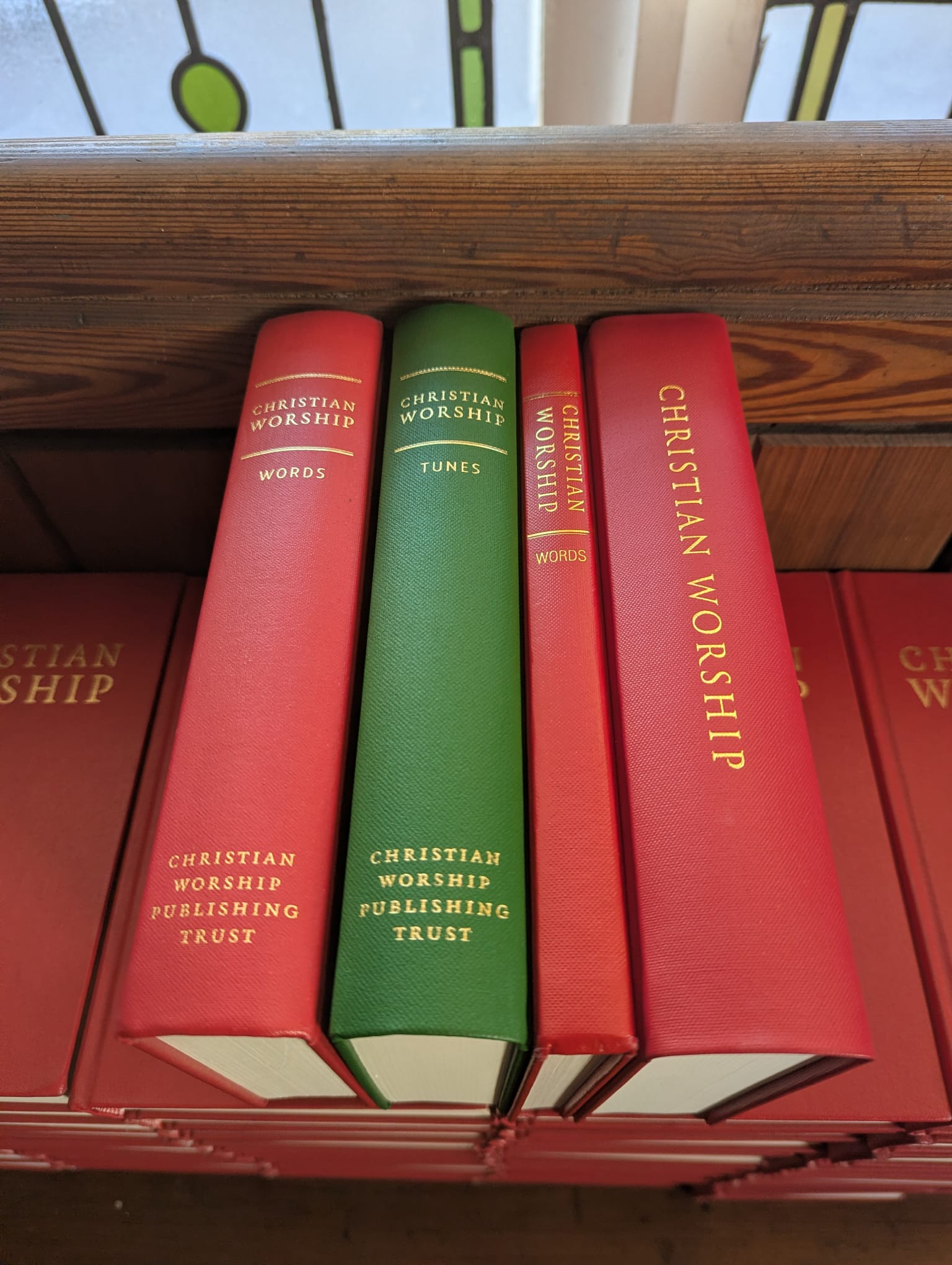 Four hymnbooks sit on their edges atop of a pile of red hymnbooks. The newest 'Combined Words and Music' edition is the widest and furthest to the right